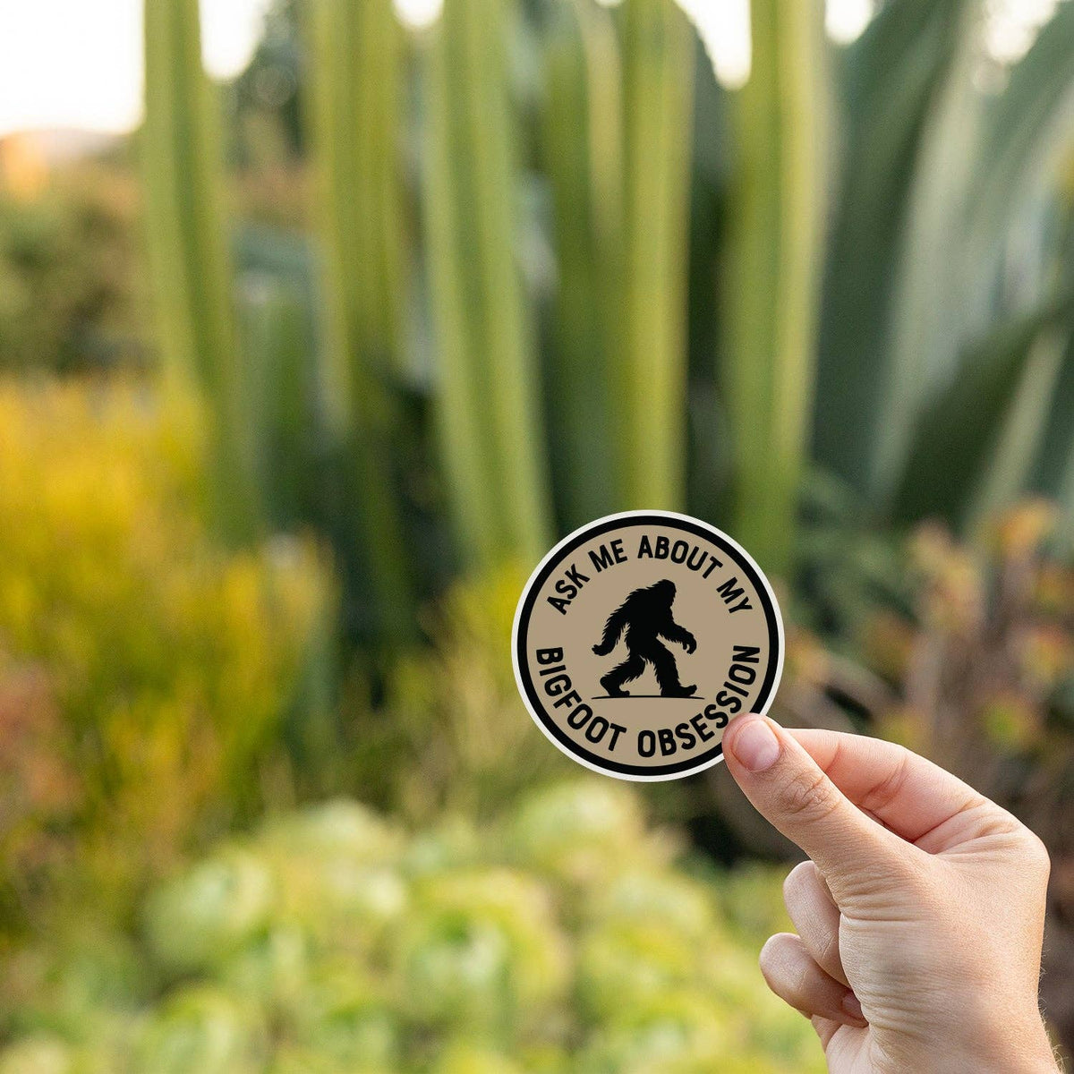Held up near tall cactus and succulents, the Ask Me About My Bigfoot Obsession sticker glows in low sun.