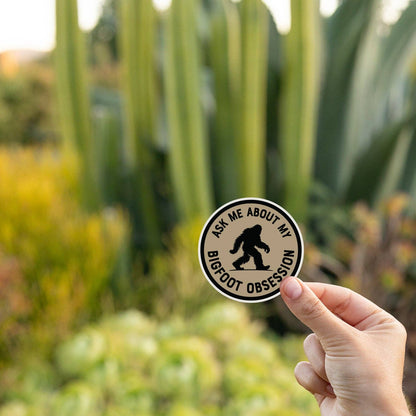 Held up near tall cactus and succulents, the Ask Me About My Bigfoot Obsession sticker glows in low sun.