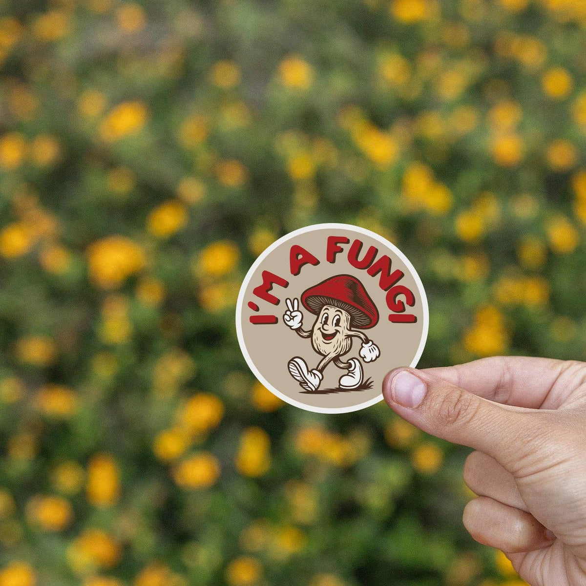 Two fingers hold the I'm a Fungi sticker before a bank of yellow wildflowers.