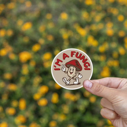 Two fingers hold the I'm a Fungi sticker before a bank of yellow wildflowers.