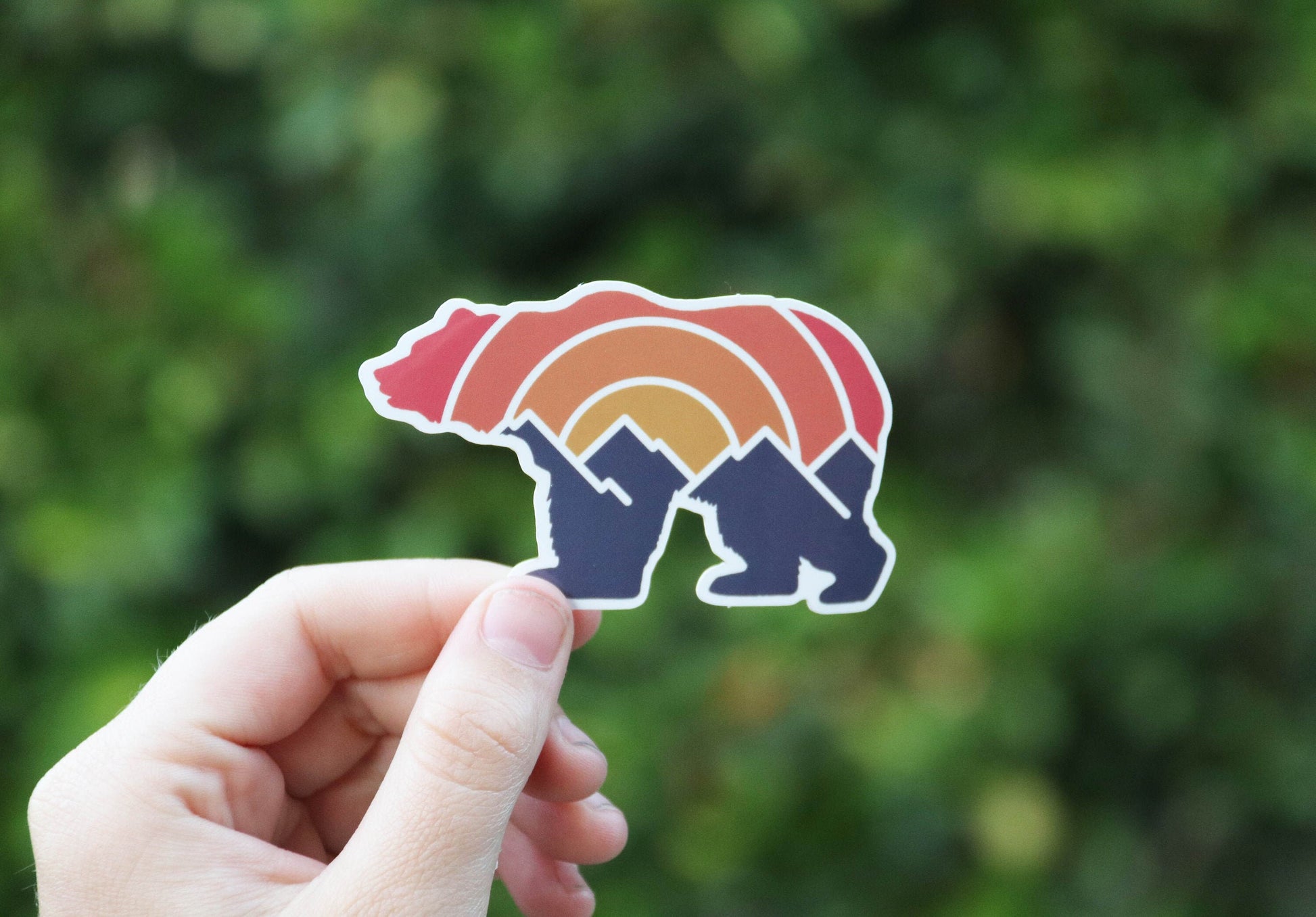 Deep green foliage fills the frame behind the Adventure Grizzly Bear sticker held in a raised hand.