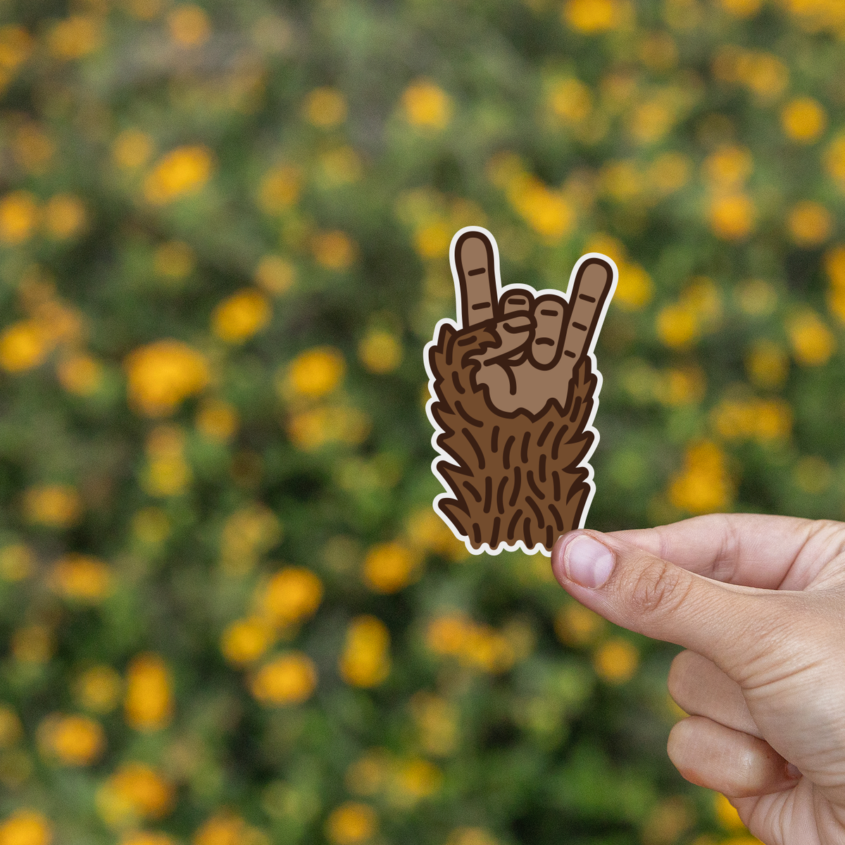 Fingers lift the Bigfoot Rocks sticker in front of a field of yellow blooms and green brush.