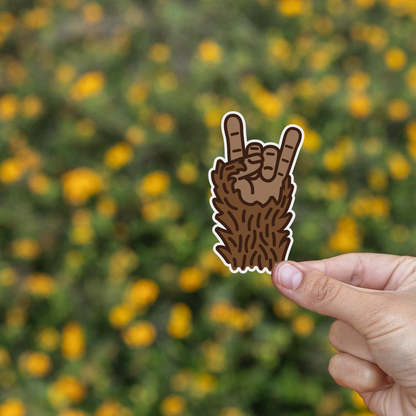Fingers lift the Bigfoot Rocks sticker in front of a field of yellow blooms and green brush.