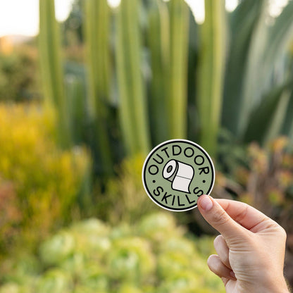 A hand holds the Outdoor Skills sticker in front of tall cactus and succulents at sunset.