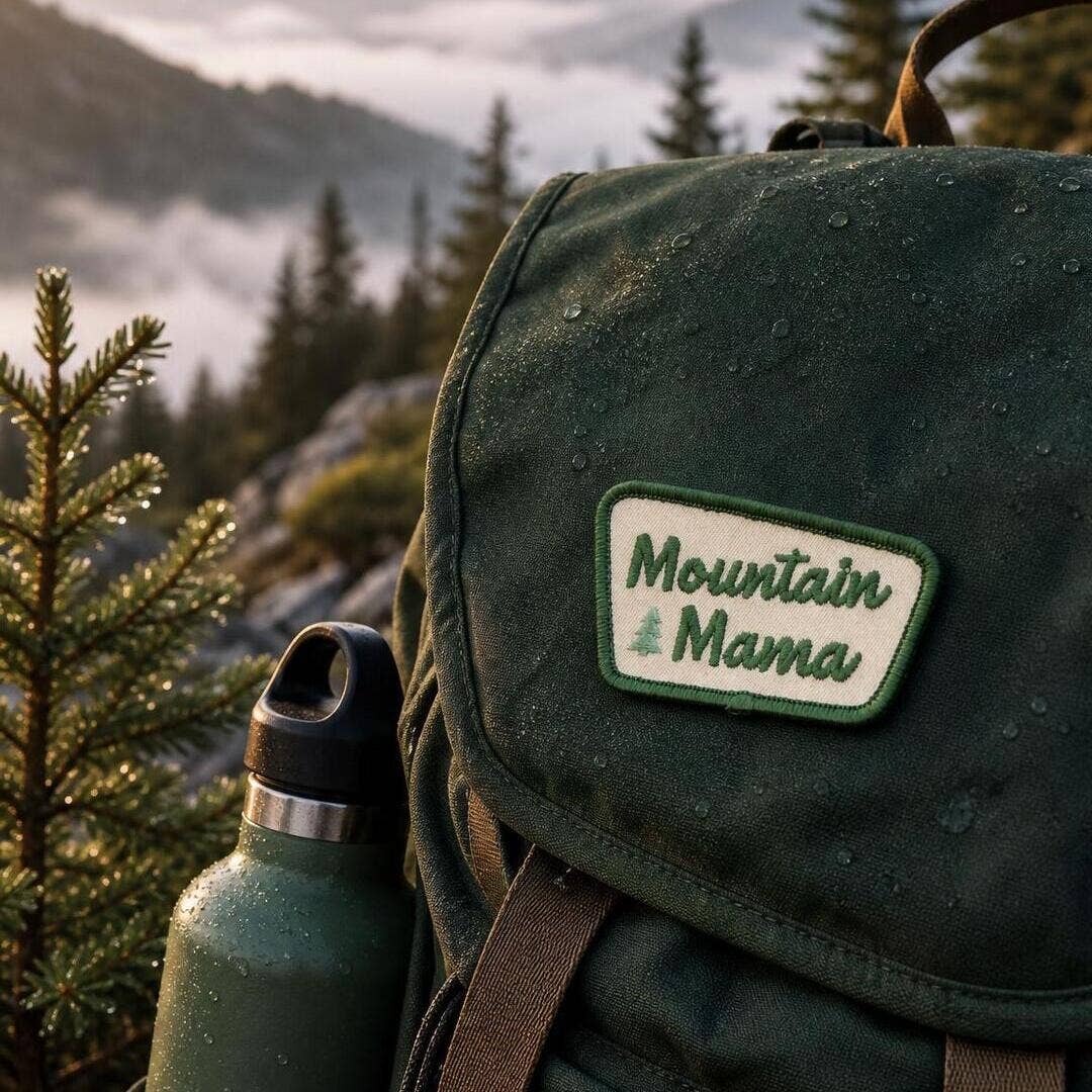 Sewn to a wet green canvas backpack, the Mountain Mama patch sits above a bottle with misty peaks behind.