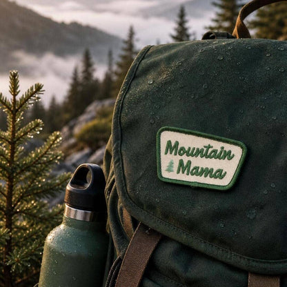 Sewn to a wet green canvas backpack, the Mountain Mama patch sits above a bottle with misty peaks behind.