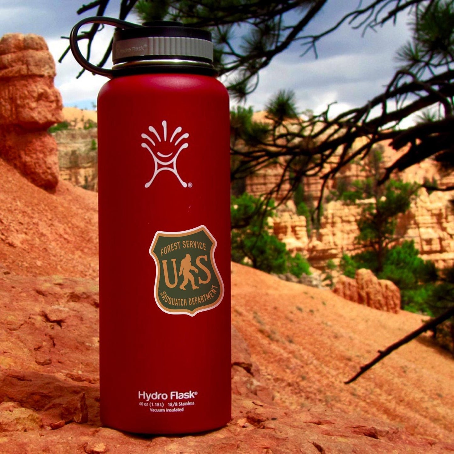 A red Hydro Flask on red rock carries the US Sasquatch Department sticker with canyon spires behind.