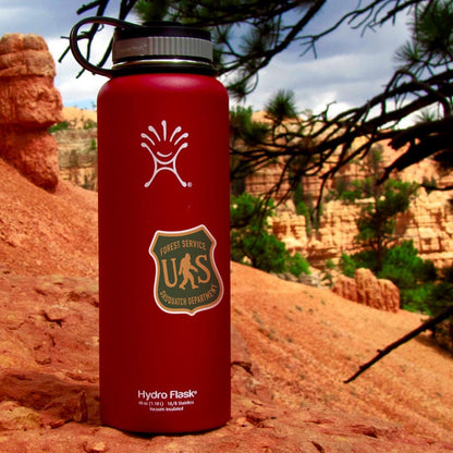 A red Hydro Flask on red rock carries the US Sasquatch Department sticker with canyon spires behind.