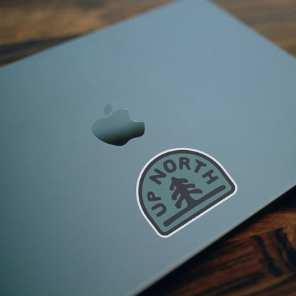 Stuck below the glowing apple on a laptop lid, the Up North sticker sits on brushed grey metal.