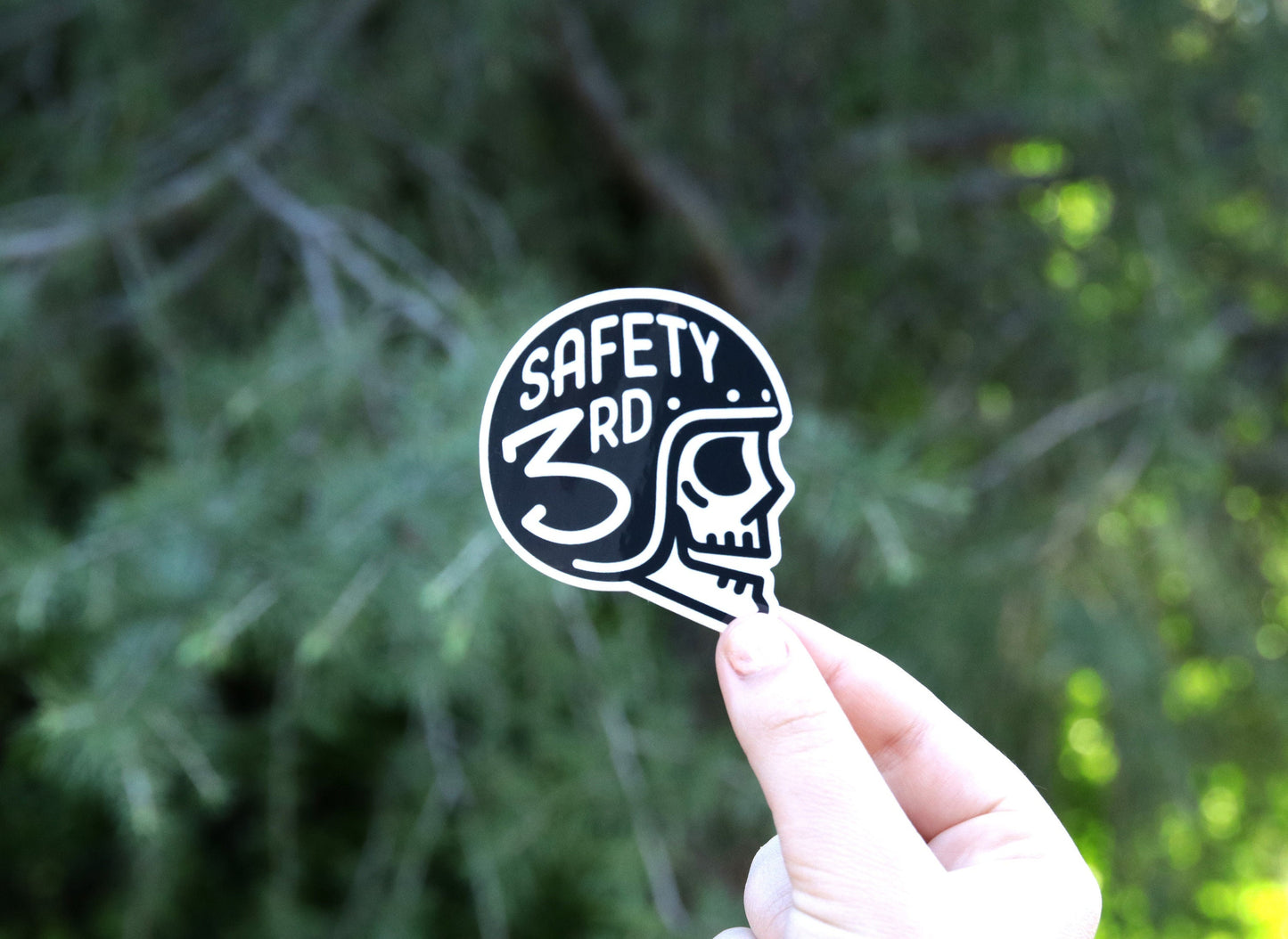 Raised toward pine branches, the Safety 3rd Helmet sticker sits between two fingers in daylight.