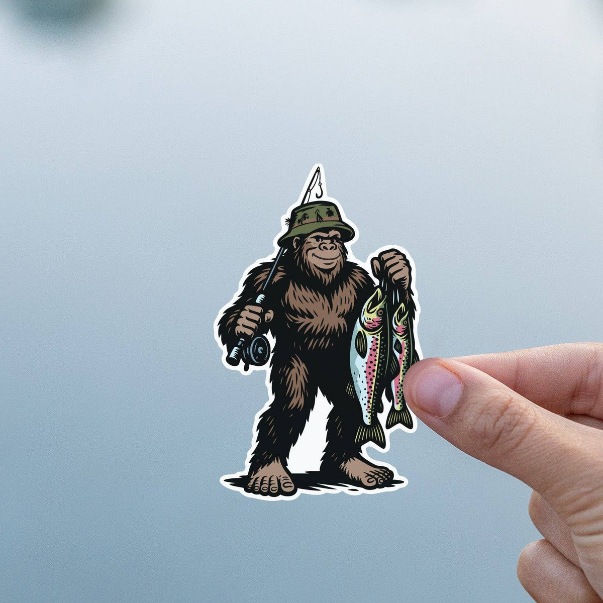 Fingers hold the Bigfoot Fishing sticker against a soft blue and grey blur of water and sky.
