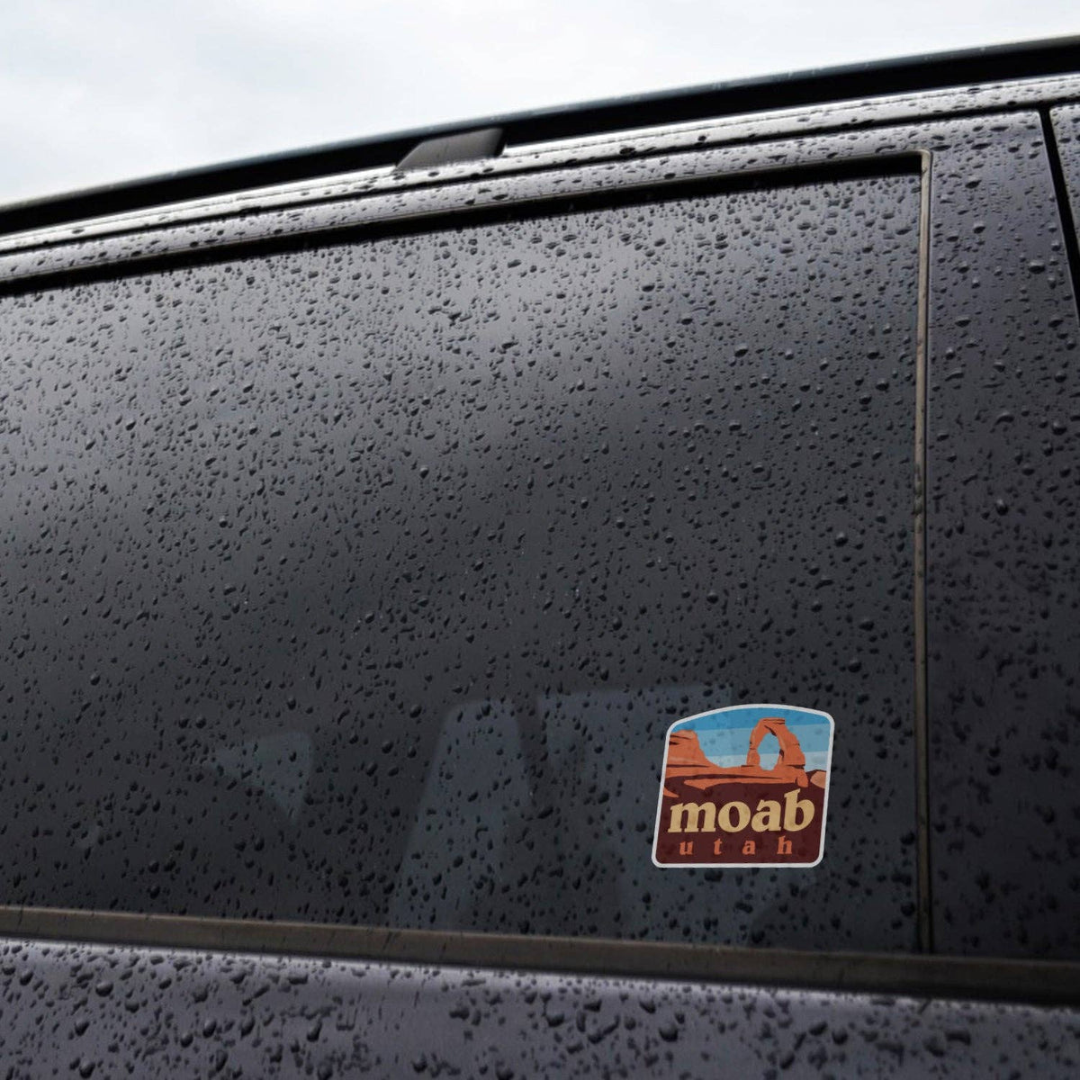 Rain beads across a dark car window where the Moab Utah sticker holds its place near the door frame.