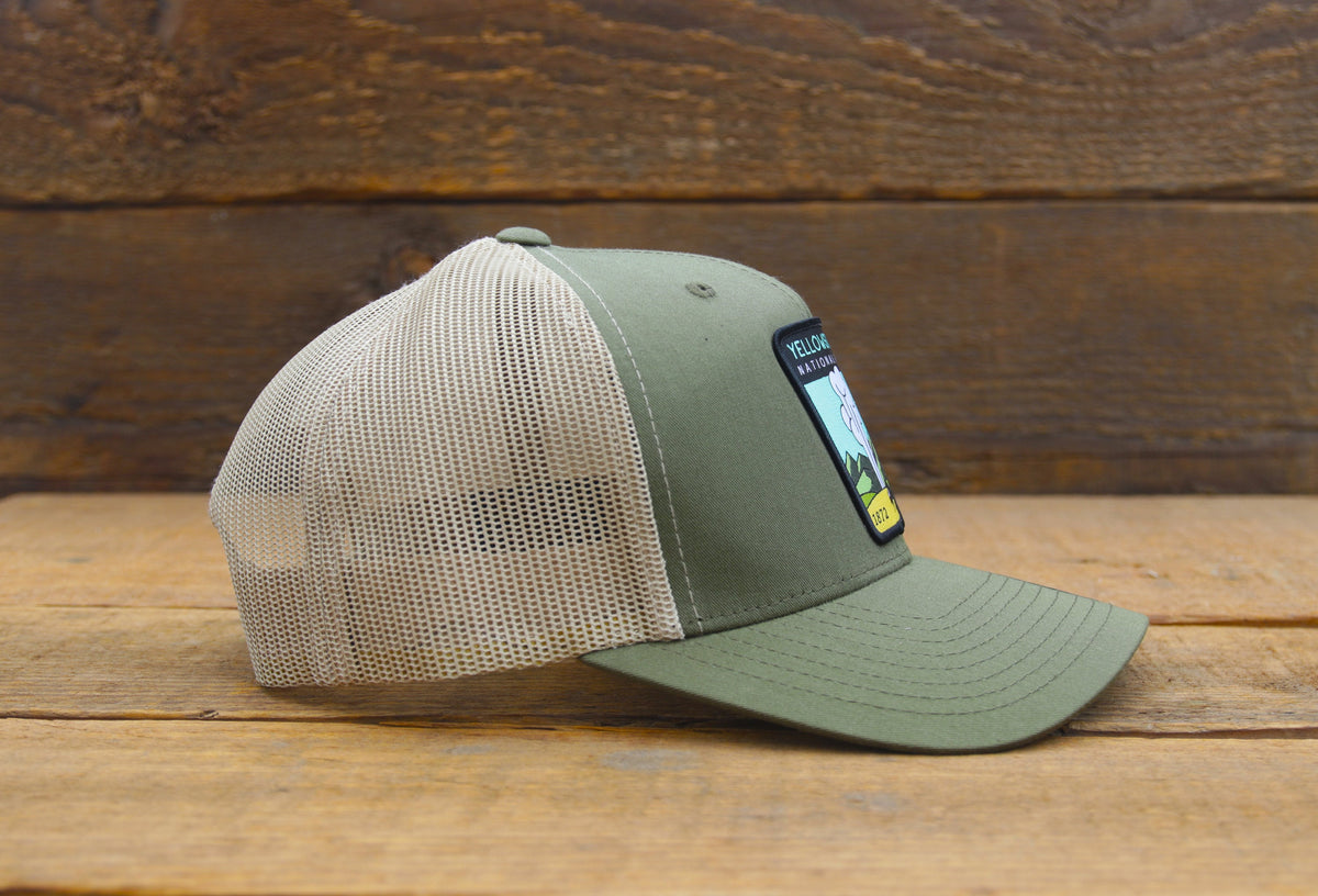 Side profile of the olive Yellowstone trucker hat, tan mesh spanning the crown above a wooden shelf.