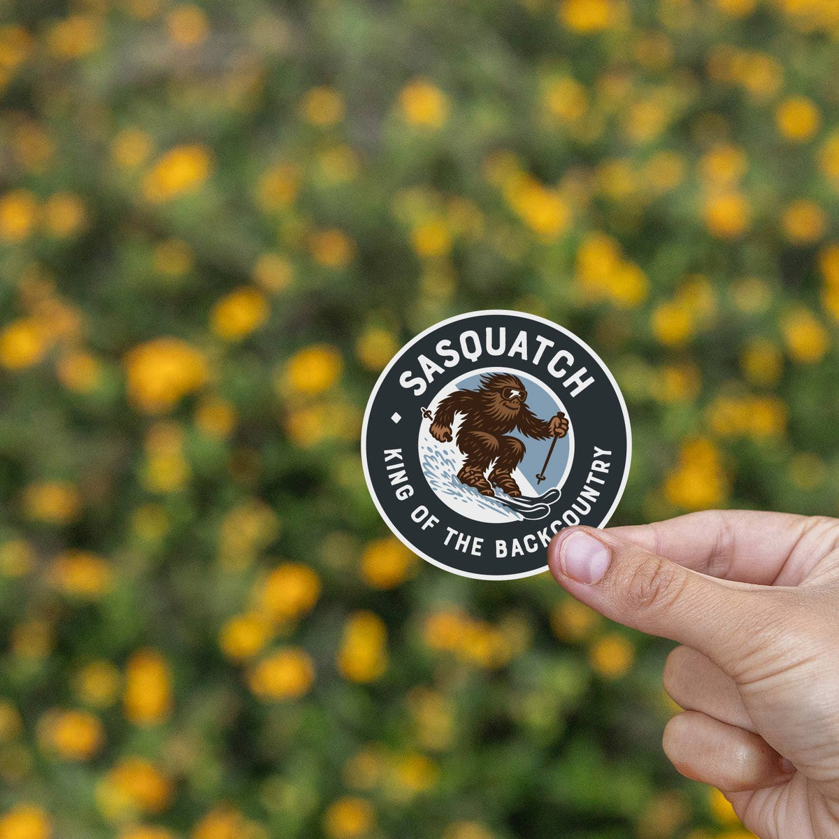 Yellow wildflowers blur behind the Sasquatch King of the Backcountry sticker held at the edge of the frame.