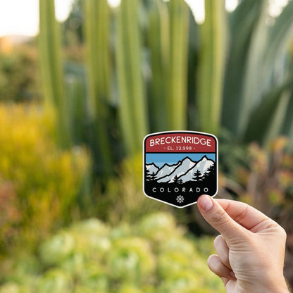 Held before tall cactus and garden greenery, the Breckenridge Colorado sticker catches late daylight.