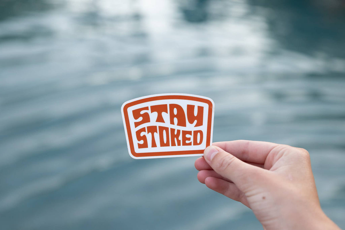A hand holds the orange Stay Stoked sticker in front of rippling water on an overcast day.