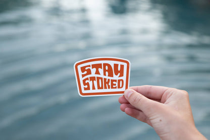 A hand holds the orange Stay Stoked sticker in front of rippling water on an overcast day.