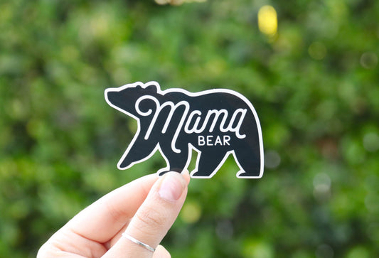 Held at arm's length, the Mama Bear sticker reads small against leafy green with a yellow bloom behind.