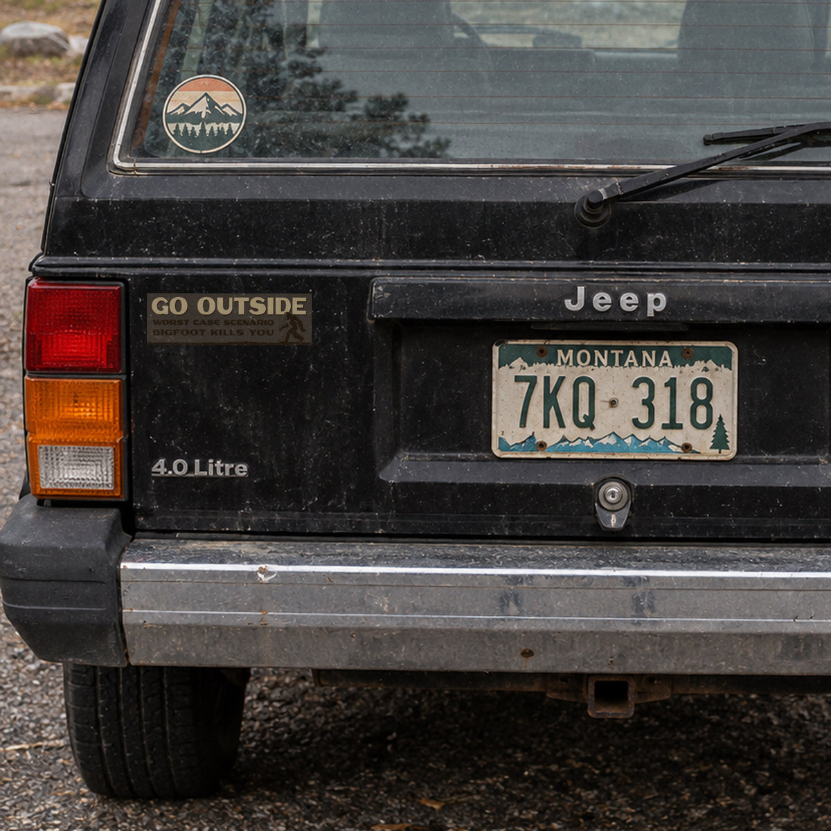 A black Jeep Cherokee's tailgate shows the Go Outside Worst Case Scenario Bigfoot Kills You sticker beside its taillight.
