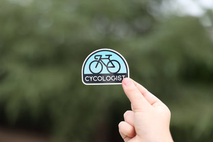 Fingertips lift the Cycologist sticker at arm's length, small against a soft wall of evergreen trees.