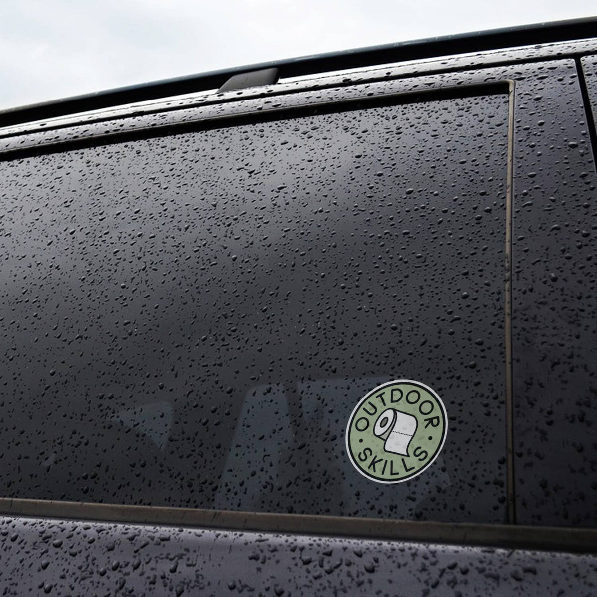 On the rain beaded window of a black car, the Outdoor Skills sticker keeps its green circle bright.