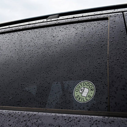On the rain beaded window of a black car, the Outdoor Skills sticker keeps its green circle bright.