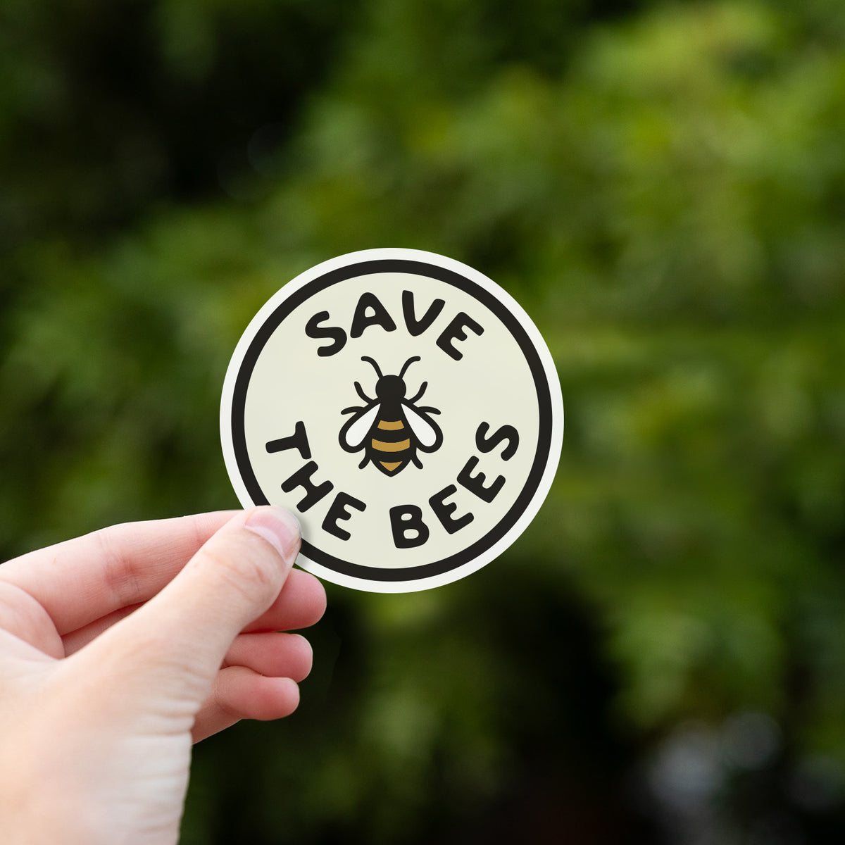 Fingers hold the Save The Bees sticker up against a backdrop of dark green leafy branches.