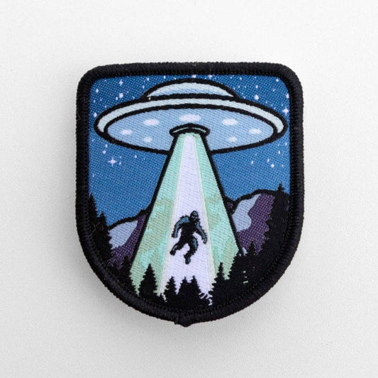 Flat on white, the Bigfoot UFO Conspiracy Patch shows a saucer beaming a sasquatch up over pine trees.