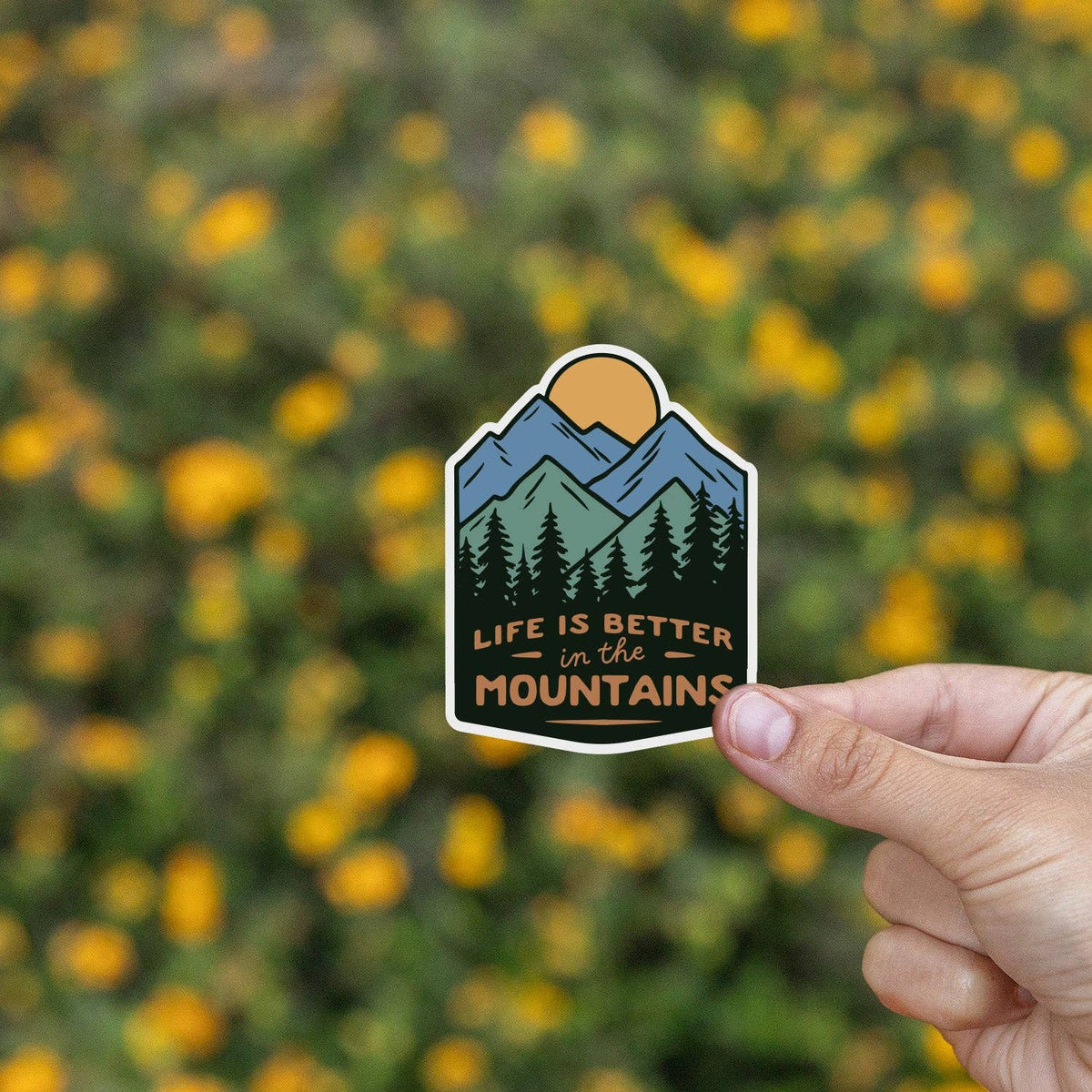 Fingers hold the Life is Better In The Mountains sticker before a blurred field of yellow wildflowers.