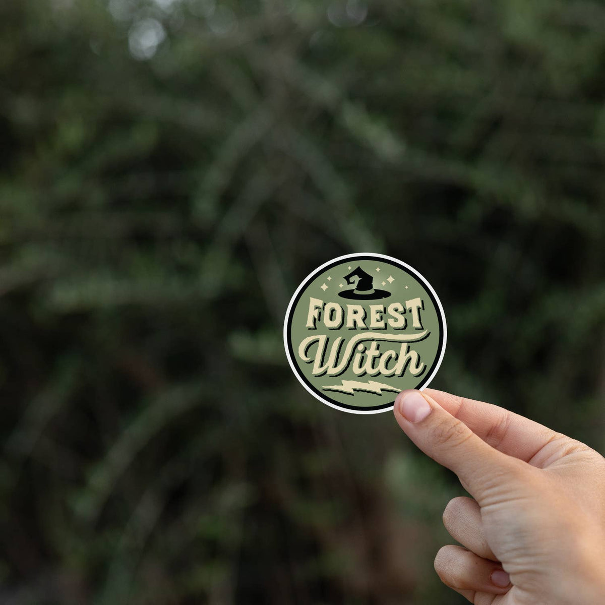Fingers hold the Forest Witch sticker in front of blurred evergreen branches.