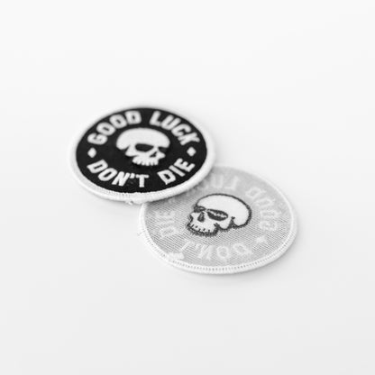 Two Good Luck Don't Die patches lean together, one showing the stitched skull front and one its pale gauzy backing.