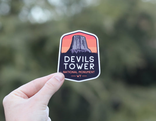 Pinched low by the thumb, the Devils Tower sticker tilts slightly with soft evergreens filling the background.