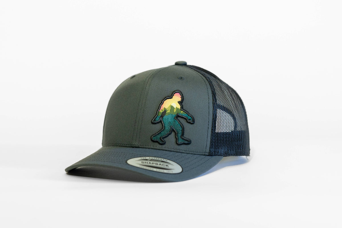 Front angle on the charcoal Bigfoot Adventure trucker hat, black mesh panels rising behind the stitched Sasquatch.