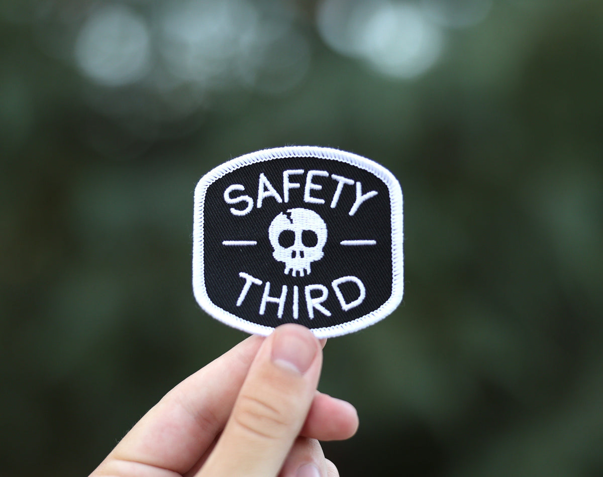 Held between finger and thumb outdoors, the Safety Third patch catches light against soft green leaves.