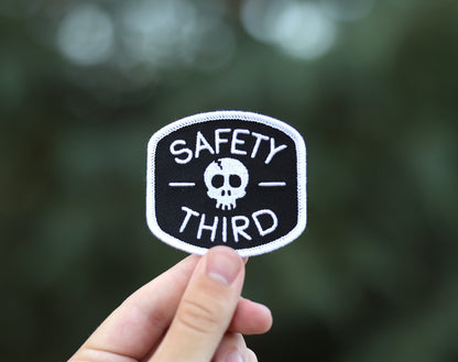 Held between finger and thumb outdoors, the Safety Third patch catches light against soft green leaves.