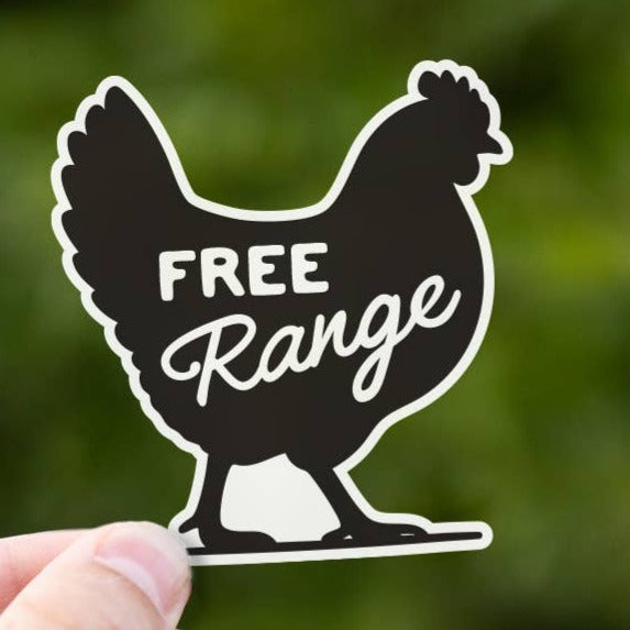 A hand holds up the black hen shaped Free Range sticker against blurred green leaves outdoors.