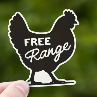 A hand holds up the black hen shaped Free Range sticker against blurred green leaves outdoors.