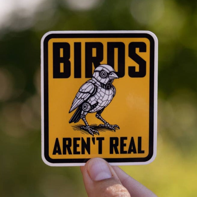 A hand holds up the yellow Birds Aren't Real sticker with its perched crow, green leaves blurred behind.