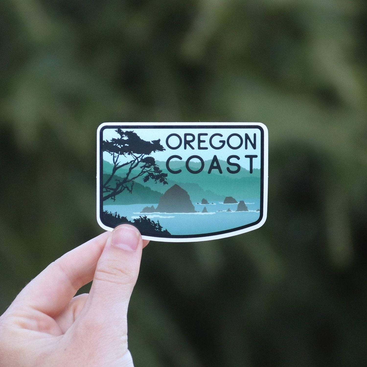 A hand holds the Oregon Coast sticker close to the lens, its sea stacks and pine branch clear against blurred green.