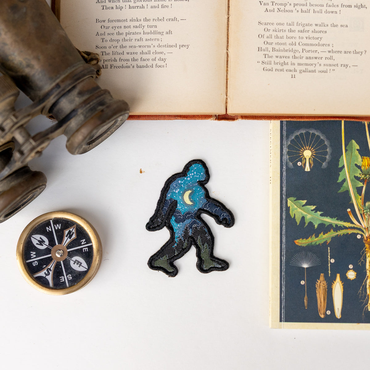 Between a compass and old binoculars, the embroidered Bigfoot Night Sky Patch rests on a page from a vintage book.