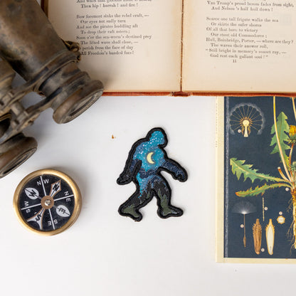 Between a compass and old binoculars, the embroidered Bigfoot Night Sky Patch rests on a page from a vintage book.