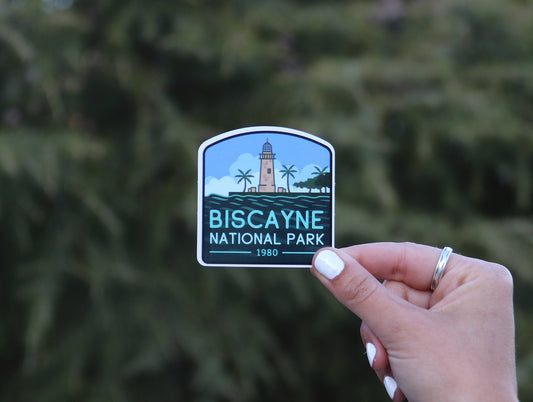 Backed by blurred green branches, the Biscayne National Park sticker sits small between finger and thumb.