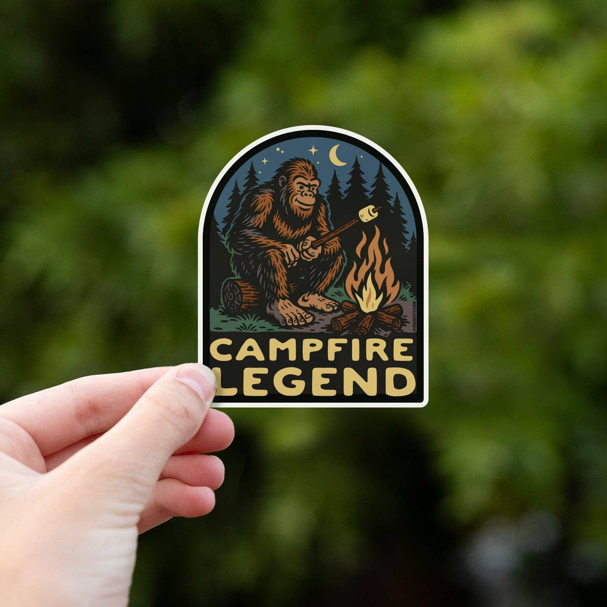 Held up against sunlit green leaves, the Campfire Legend sticker shows its gold lettering and orange flames.