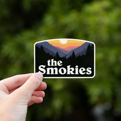 Fingers hold the The Smokies sticker out in front of soft green foliage lit by daylight.