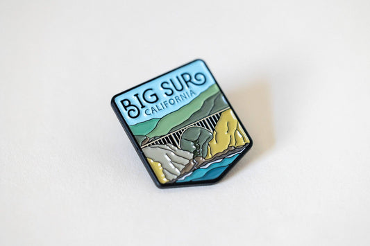 Tilted on a plain white surface, the Big Sur California pin catches light along its black metal edges.