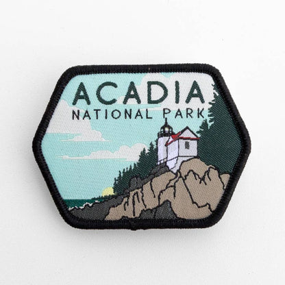 Flat on a white surface, the Acadia National Park patch shows a lighthouse on rocky cliffs above the water.