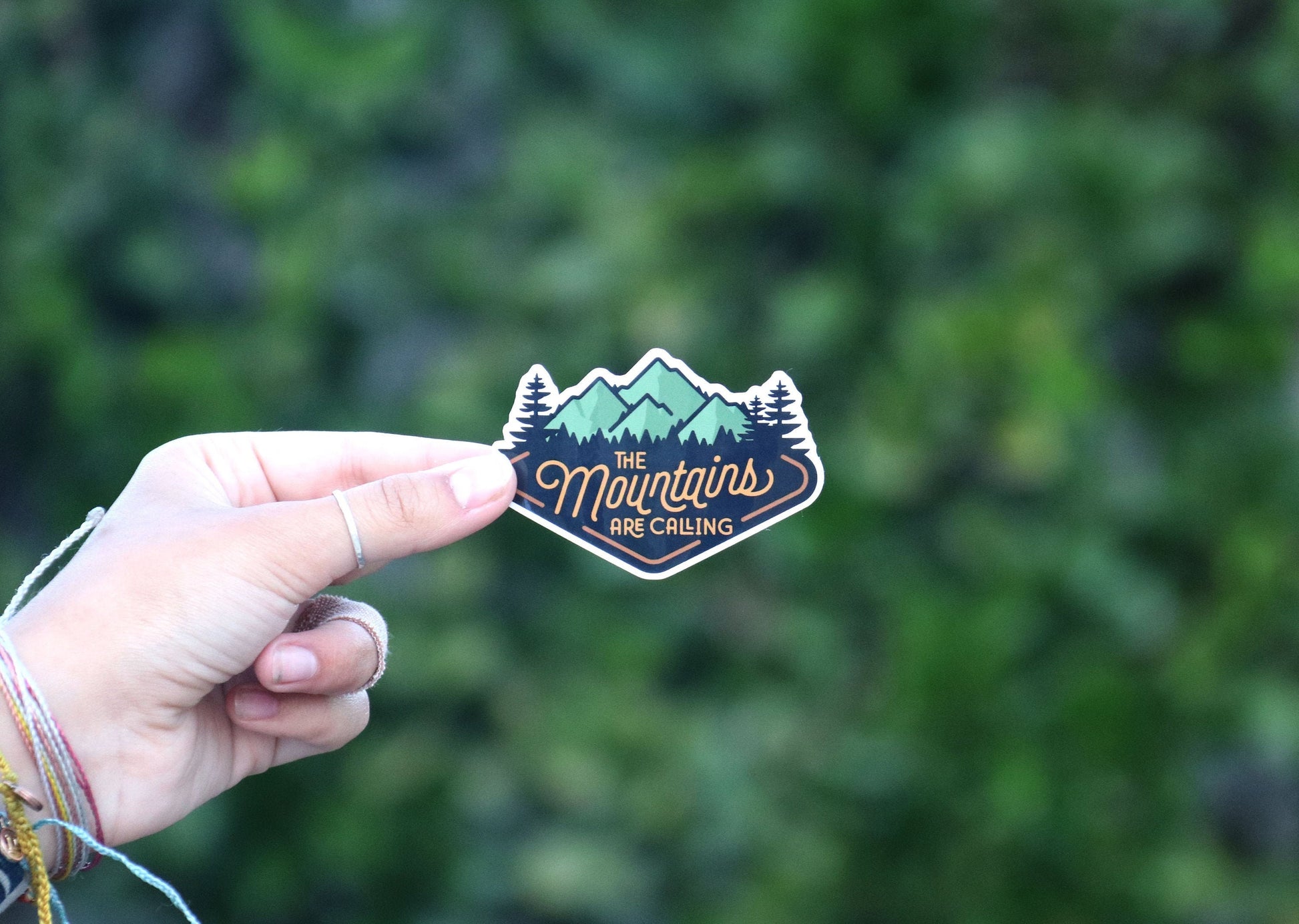 Bracelet strings dangle as a hand holds The Mountains Are Calling sticker before a wall of shrubs.