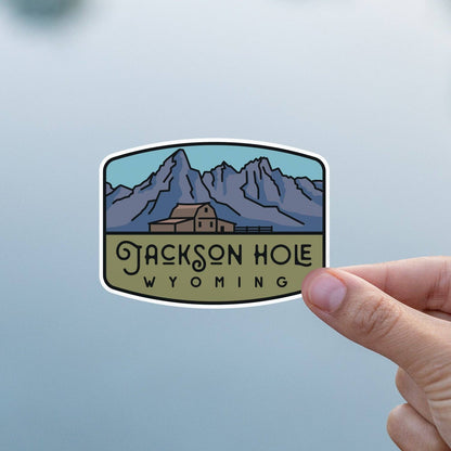 Held against pale grey haze, the Jackson Hole Wyoming sticker shows its barn, fence line and mountain ridge.