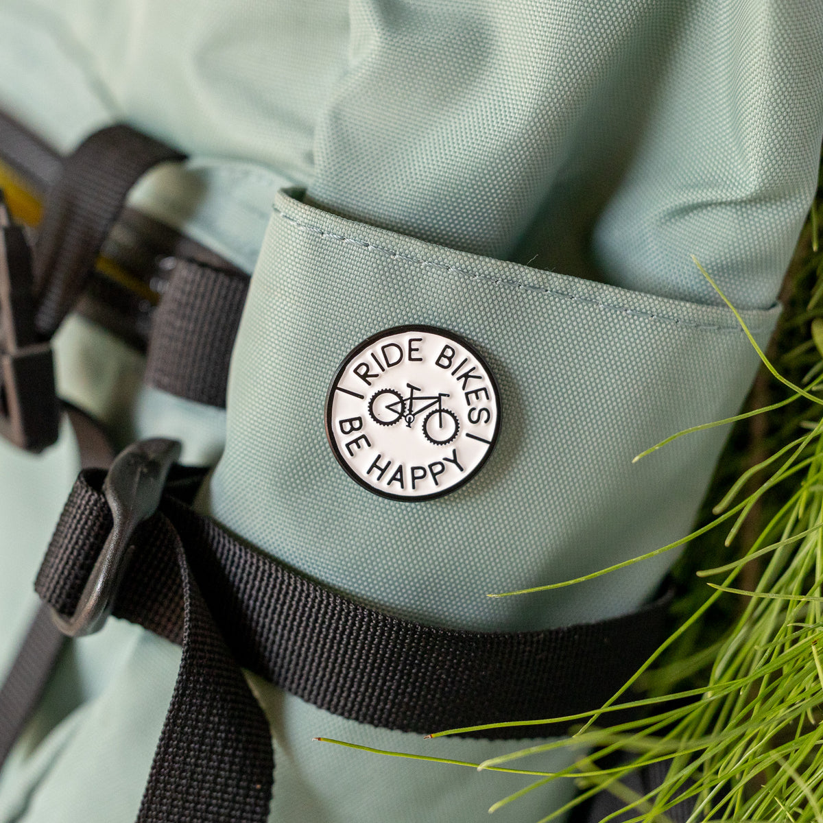Clipped to a sage green bag strap, the Ride Bikes Be Happy pin rests near blades of grass and a buckle.