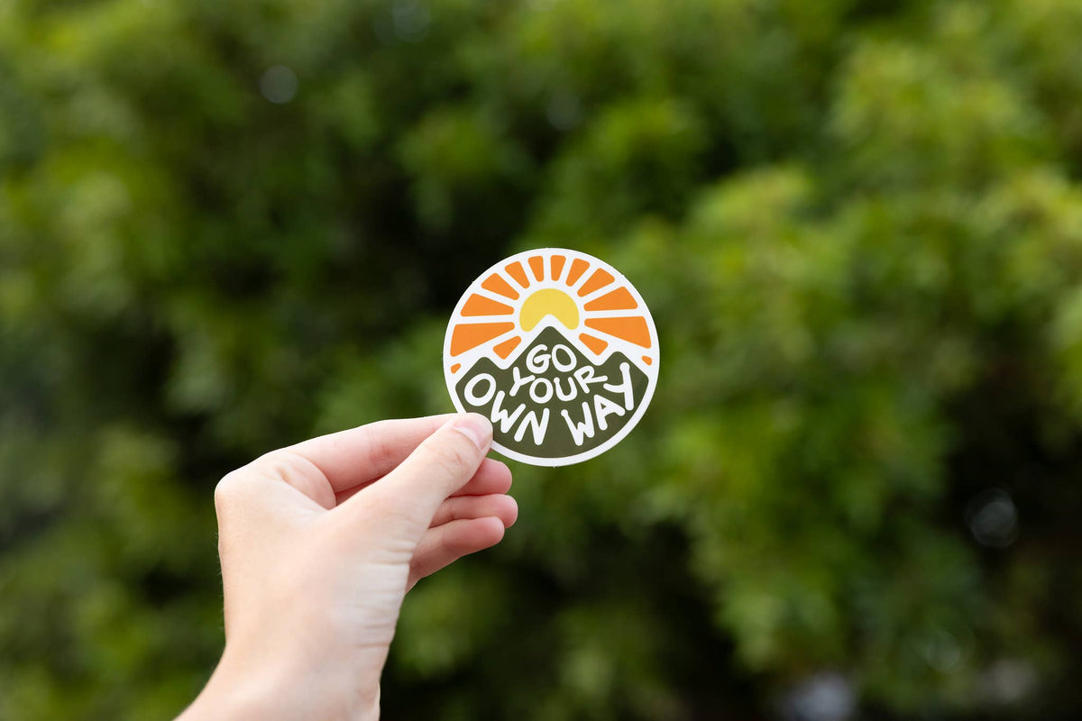 Held out at arm's length, the Go Your Own Way sticker catches light against dark green foliage.